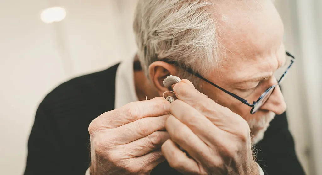 An older man with grey hair holding a hearing aid and inspecting it as if trying to figure out how to troubleshoot common hearing aid issues